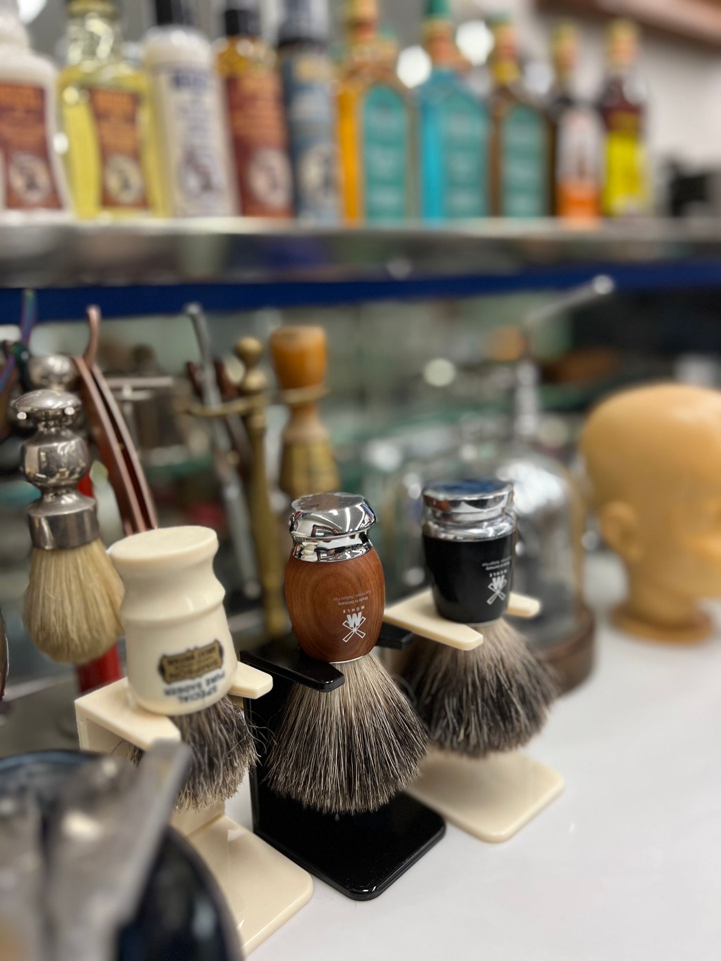 Shaving brushes in stands with bottles of grooming products on a shelf in the background.