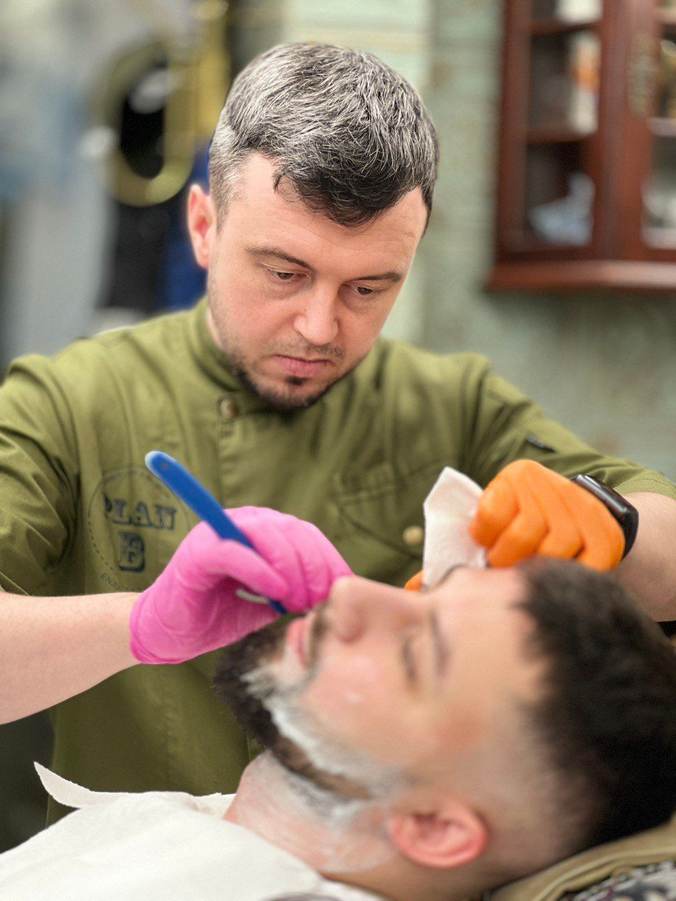 Barber using razor to shape client's beard, wearing gloves in barbershop.